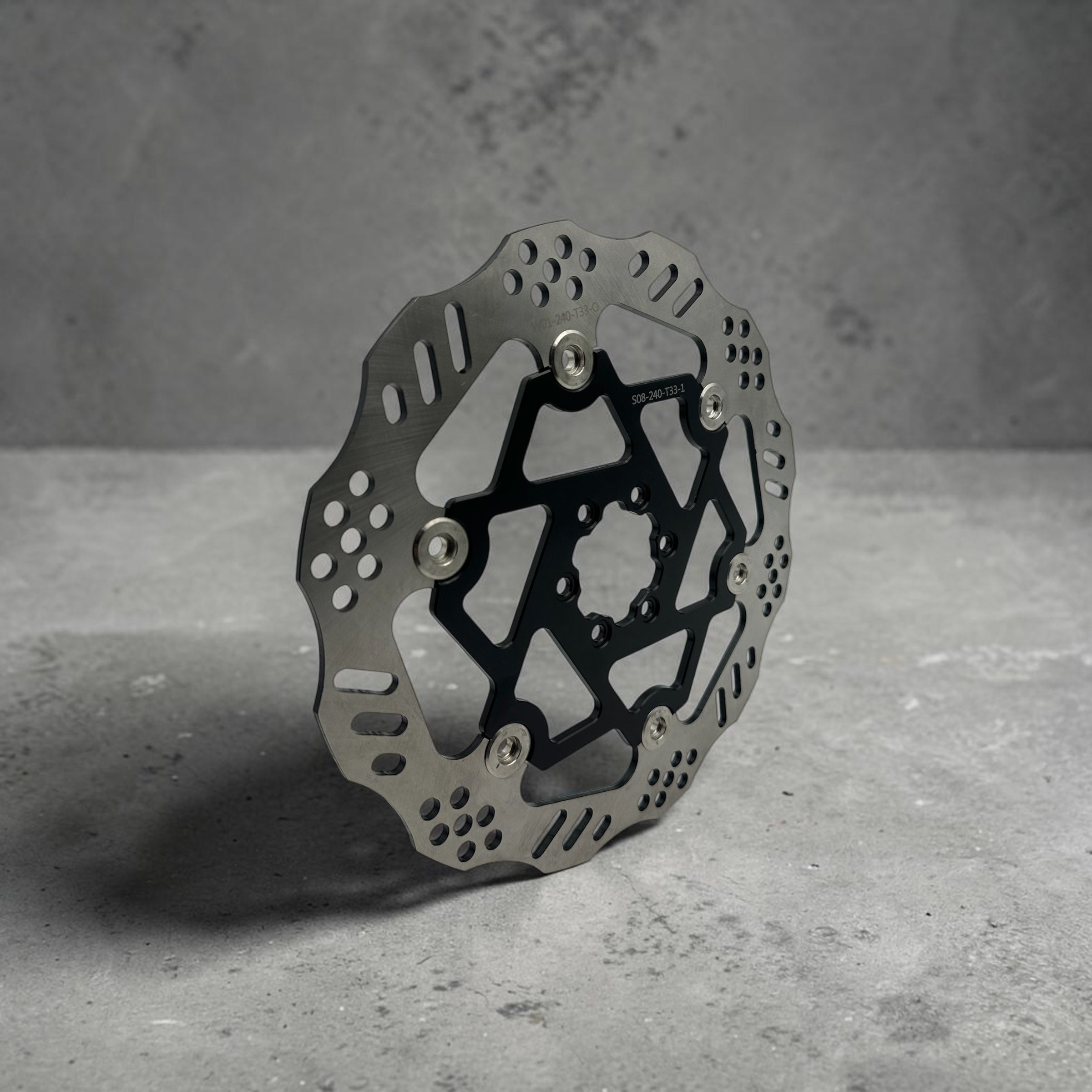 A metallic bicycle disc brake rotor with a black center core and silver mounting hardware, photographed on a textured gray concrete 240mm floating ultra bee brake rotor