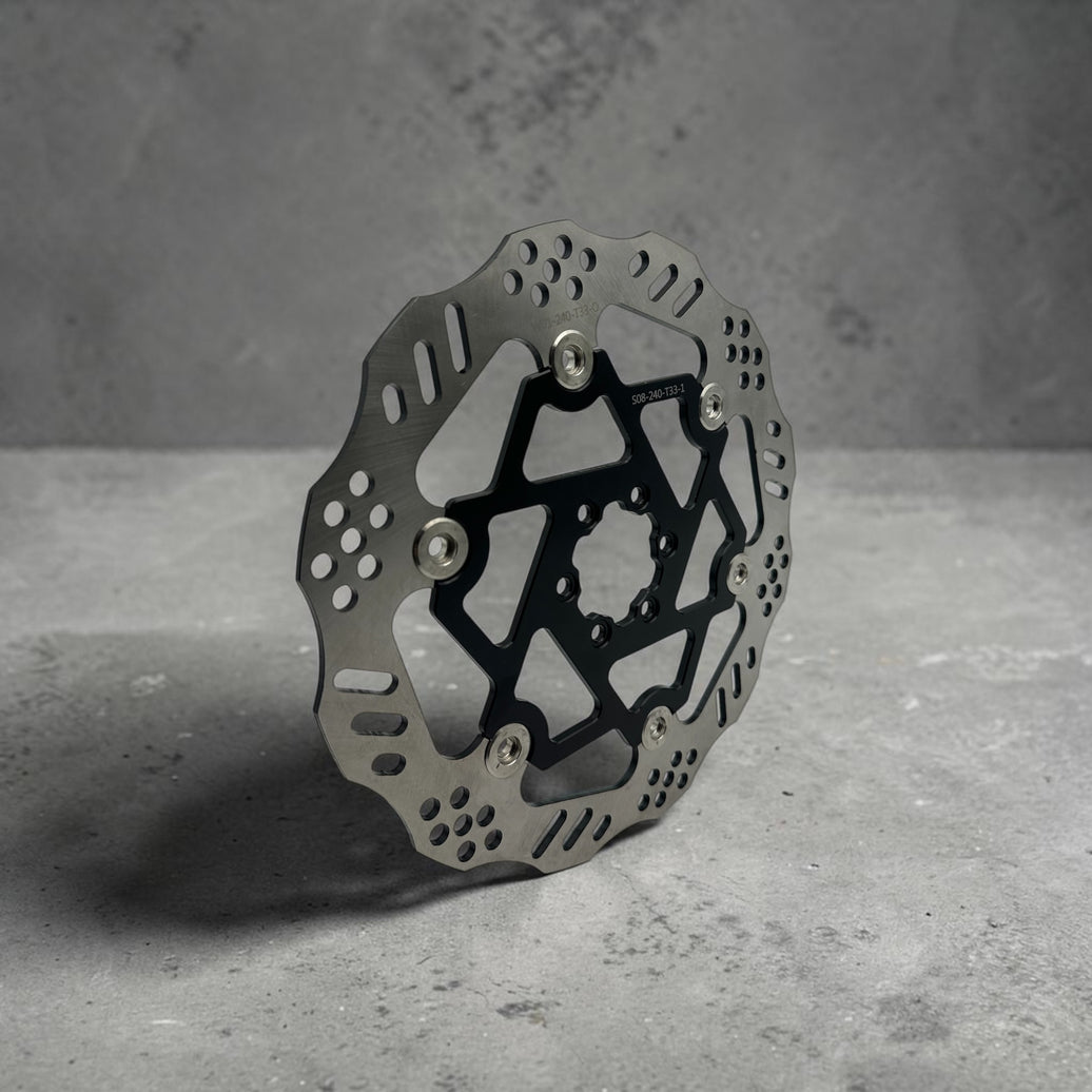 A metallic bicycle disc brake rotor with a black center core and silver mounting hardware, photographed on a textured gray concrete 240mm floating ultra bee brake rotor