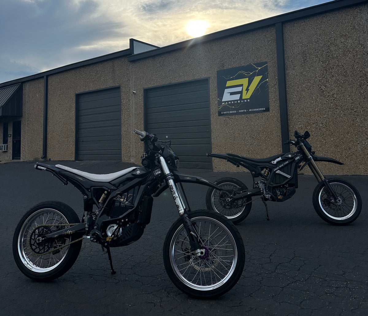 Two black electric dirt bikes parked outdoors in front of a tan building with garage doors and a sign that reads EV Warehouse. The photo is taken at sunset with the sun low on the horizon and clouds in the blue sky.